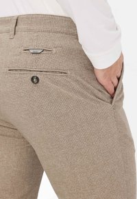Beige geruite broek met een gestructureerde stof, voorzien van een achterzak met een zwarte knop en een klein merkembleem boven de zak.
