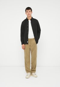 Svart knappopp skjorte over en hvit t-skjorte, kombinert med beige kordfløyelsbukser og hvite joggesko. Modellen står mot en ensfarget bakgrunn.