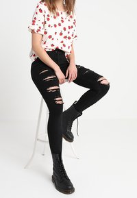 Blusa blanca con estampado floral rojo, jeans skinny negros rotos y botas negras con cordones, posicionados sobre un taburete blanco contra un fondo claro.