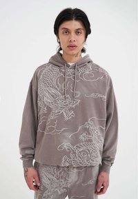 Felpa grigia con design di drago in ricamo bianco, caratterizzata da una vestibilità ampia, cappuccio con cordino e polsini a coste. Pantaloni semplici in tessuto coordinato.