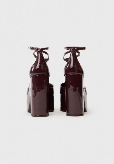 Scarpe con tacco alto in pelle verniciata color bordeaux, dotate di un spesso tacco a blocco, cinturini alla caviglia e una finitura liscia e lucida. La vista sul retro rivela un design a taglio.