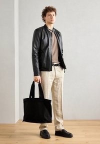 Bolso de tela negro con asas resistentes, combinado con una chaqueta de cuero negra, pantalones de lino beige y zapatos negros sin cordones.
