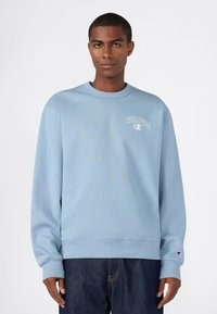Joven con sudadera ligera azul de Champion y jeans oscuros, de pie frente a un fondo blanco, mirando hacia adelante.