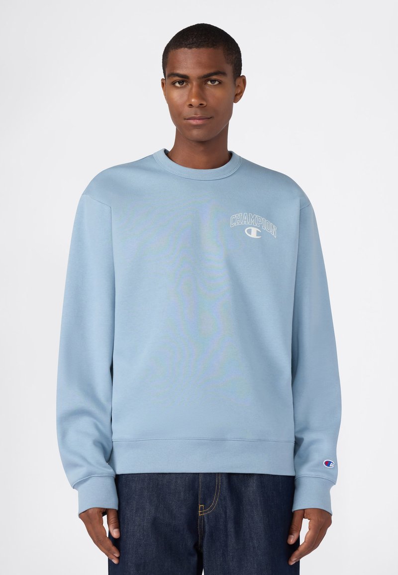 Giovane uomo che indossa un pullover Champion azzurro chiaro e jeans blu scuro, in piedi contro uno sfondo bianco, rivolto in avanti.