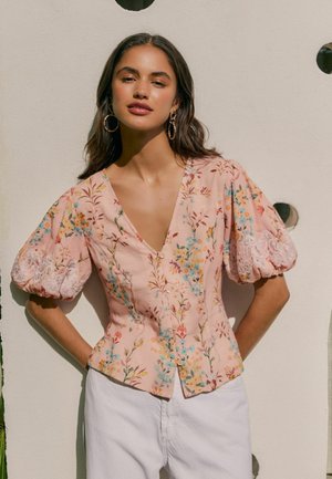 Jonge vrouw met lang donker haar, grote ronde oorbellen en een roze bloemenblouse met pofmouwen en een witte broek, staand tegen een muur.