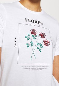 Hvid bomulds T-shirt med et grafisk motiv af tre røde roser med grønne blade, kantet tekst og glitrende udsmykninger.