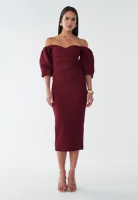 Abito borgogna off-the-shoulder con maniche a sbuffo, corpetto aderente, gonna al ginocchio e tessuto testurizzato. Sandali dorati con cinturini completano il look.