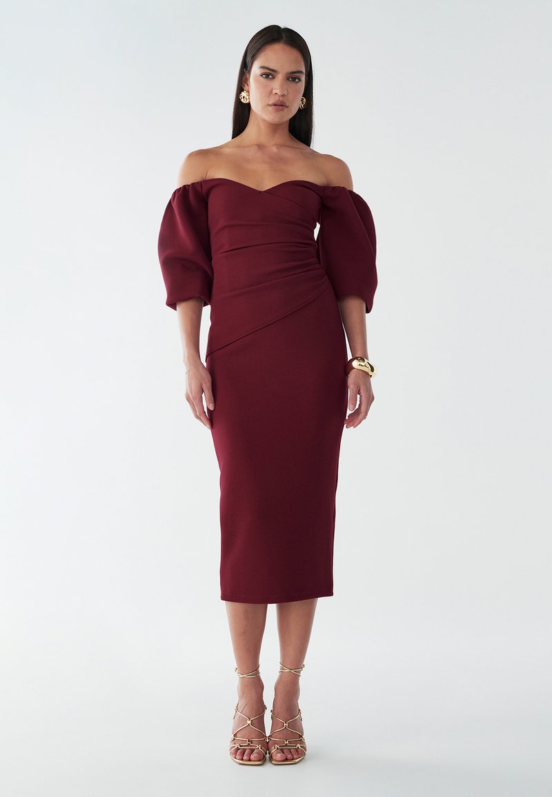 Abito borgogna off-the-shoulder con maniche a sbuffo, corpetto aderente, gonna al ginocchio e tessuto testurizzato. Sandali dorati con cinturini completano il look.