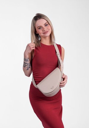 Krosbodová taška z měkké béžové kůže s zakřiveným tvarem, nastavitelným popruhem a embosovaným logem. Modelka má na sobě těsné červené šaty.