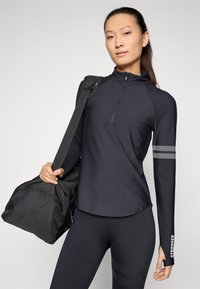 Black athletic long-sleeve top with a quarter zip, featuring two gray stripes on the left arm. Model holds a black duffel bag.