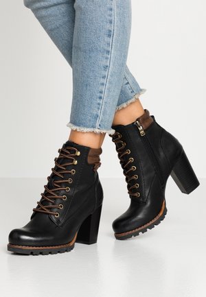 Schwarze Leder-Stiefeletten mit hohen Absätzen, braunen Schnürsenkeln und Verzierungen, getragen mit hellblauen Jeans mit ausgefranstem Saum bei gekreuzten Beinen.