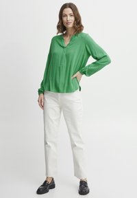 Blusa verde a maniche lunghe con colletto alla cinese, vestibilità rilassata e polsini arricciati, abbinata a pantaloni bianchi a gamba dritta e mocassini neri.