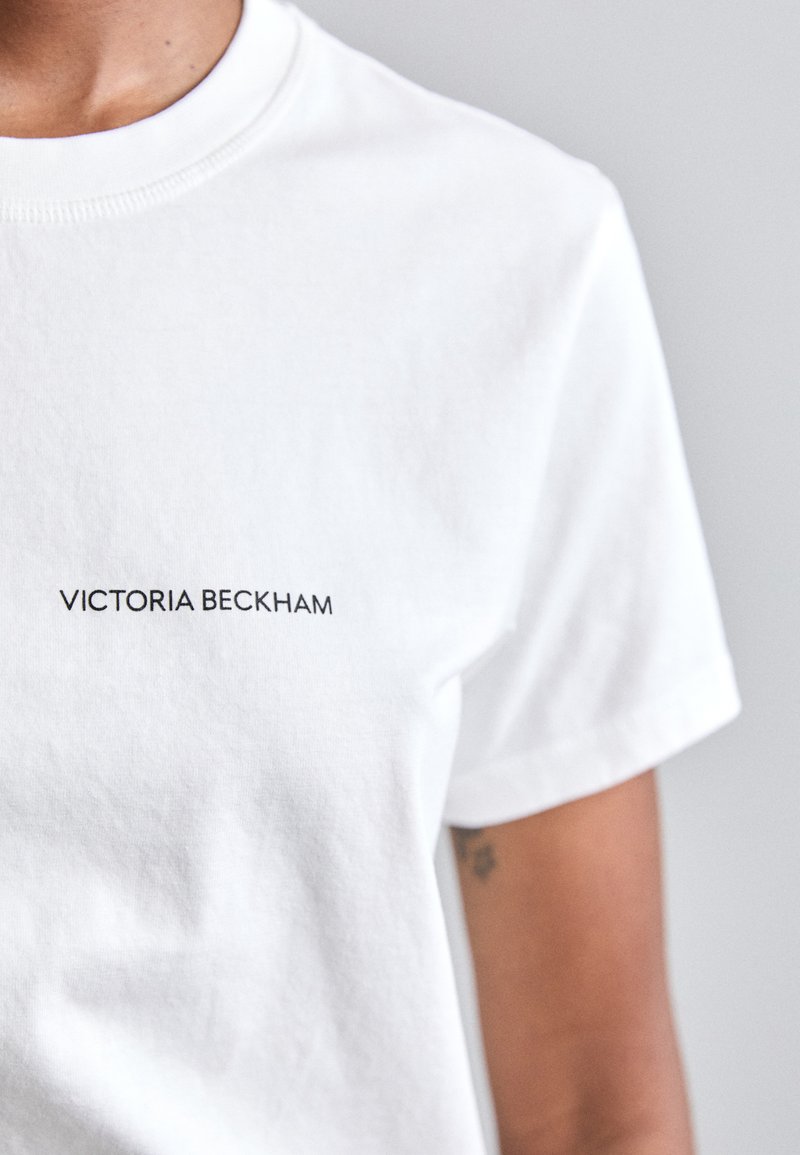Camiseta de algodón blanca con cuello redondo clásico, mangas cortas y un logo impreso "VICTORIA BECKHAM" en negro en la parte frontal.