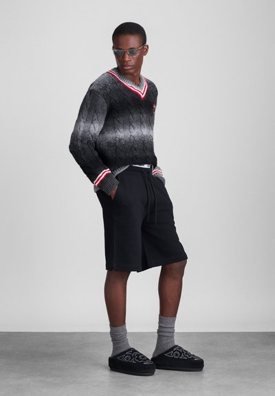Vernähteter schwarzer und grauer Pullover mit Kabelstruktur und roten Akzenten, kombiniert mit schwarzen Shorts, grauen Socken und schwarzen gemusterten Hausschuhen.
