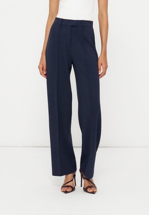 Femme portant un pantalon large bleu marine associé à des sandales noires à talons hauts et à brides, ainsi qu'un haut blanc sans manches.