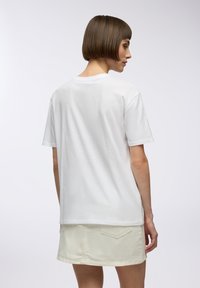 T-shirt bianco in cotone con maniche corte, taglio dritto e collo rotondo. Tessuto texturizzato con una finitura liscia. Indossato con una gonna chiara.