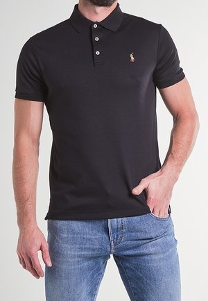 Mann trägt ein schwarzes Poloshirt mit kurzen Ärmeln und kleinem besticktem Logo sowie blaue Jeans, Hand teilweise in der Tasche.