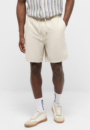 Mann trägt beige Shorts mit Kordelzug, weiße Sneaker, weiße Socken mit dem Schriftzug „MUSTANG“ und ein gestreiftes Hemd mit Knöpfen, die Hände in den Taschen.