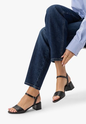 Persoană purtând blugi bleumarin și o cămașă albastru deschis ajustează sandale negre cu toc bloc, cu decupaj la vârf și curele la gleznă, în timp ce stă așezată.