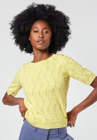 Gelbes gestricktes Kurzarm-Sweatshirt mit Wellenmuster, strukturierten Punkten sowie geripptem Ausschnitt und Saum. Kombiniert mit hellblauen Hosen.