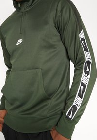 Pullover Nike verde scuro con mezza zip, dotato di tasca frontale e dettagli in bianco e nero del logo lungo la manica. Texture liscia.