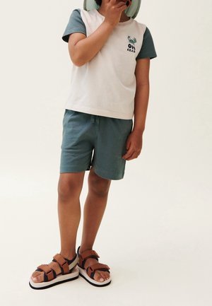 Enfant en short vert et chemise blanche aux manches vertes, portant des sandales marron et tenant la main près de la bouche, debout sur un fond clair.