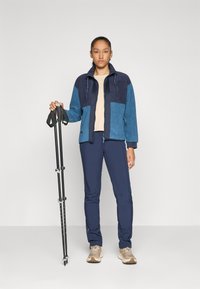 Kvinne som står i blå og marineblå fleecejakke, marineblå bukser, beige joggesko, og holder sammenbrettede svarte turstaver mot ensfarget bakgrunn.