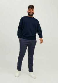 Mornarsko pleten pulover z okrolim izrezom, kombiniran s temno modrimi ožjimi hlačami in belimi supergami. Model stoji v sproščenem, priložnostnem položaju.