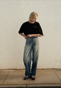 Svart oversized t-skjorte med lilla tekst, falmete blå vide jeans, og svarte spisse sko med sølvdetaljer, mot en teksturert hvit vegg.