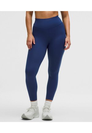 Kvinde iført marineblå leggings med højt liv, hvide crew-strømper og hvide sportssko, stående mod en ensartet lys baggrund.