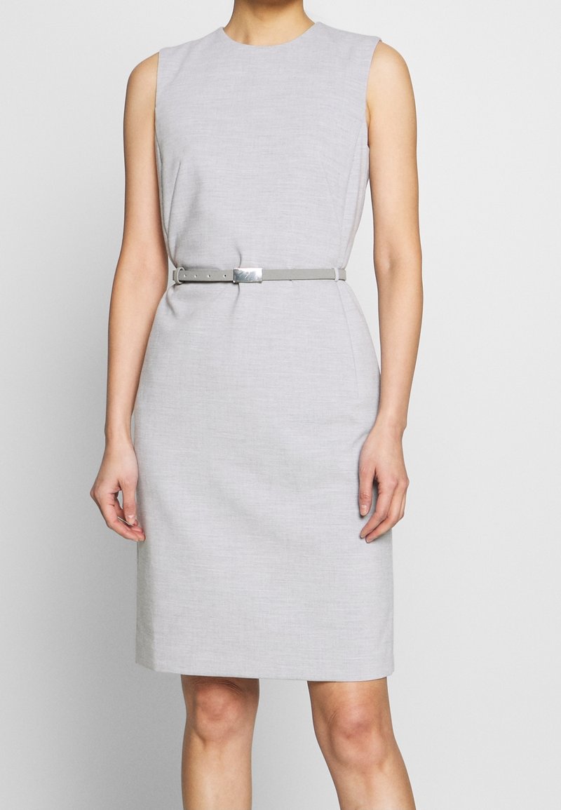 Robe sans manches gris clair longueur genoux avec une fine ceinture assortie et une boucle argentée rectangulaire portée par une personne debout.