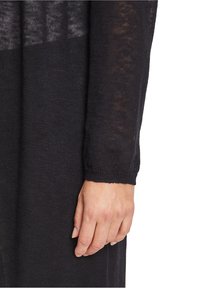 Černý pletený svetr s dlouhými rukávy, který se vyznačuje texturovaným povrchem a přiléhavým manžetou, ukazuje část ruky odpočívající na rukávu.