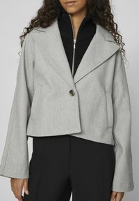 Zkrácená šedá vlněná sako s širokými klopami, uzavírání na jeden knoflík a kapsami s klopami, kombinované s černým topem s vysokým límcem pod tím.