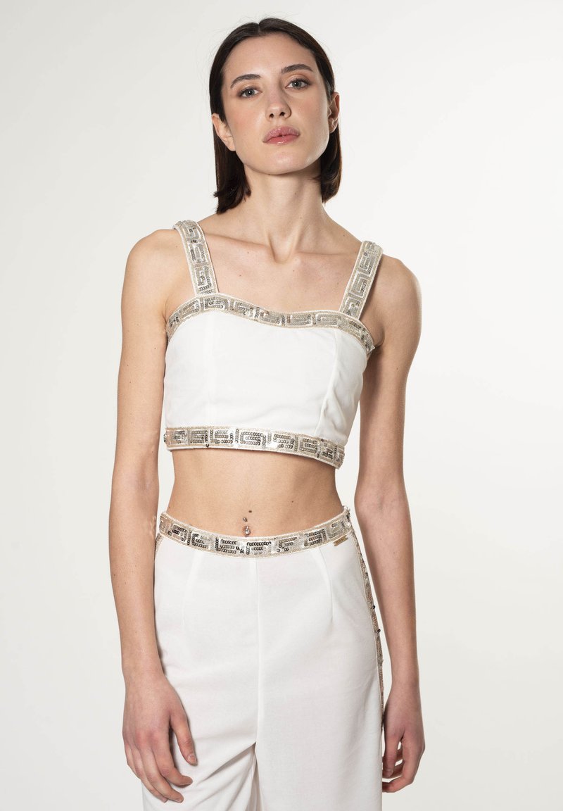 Top corto blanco con ribete de patrón geométrico plateado en los hombros y en el dobladillo. La modelo lleva pantalones de talle alto a juego, también detallados con ribete.