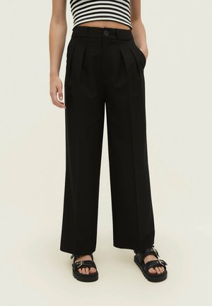 Person trägt hoch taillierte schwarze weite Hosen, schwarze Schnallensandalen und ein schwarz-weiß gestreiftes kurzes Oberteil, steht mit den Händen in den Taschen.