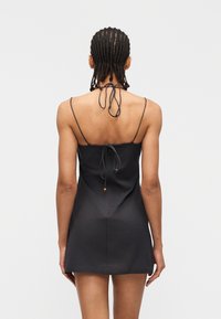 Γυναίκα με πλεξούδα που φοράει κοντό μαύρο φόρεμα με λεπτές τιράντες και δέσιμο στο πίσω μέρος, στέκεται μπροστά σε απλό γκρι φόντο.