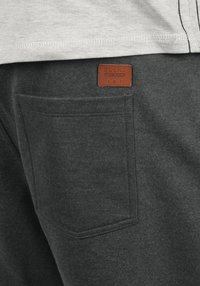 Pantalon de survêtement gris avec une poche arrière, présentant un patch en cuir marron avec l'inscription "BLEND" et des détails brodés le long des bordures.