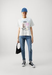 T-shirt graphique blanc avec du texte coloré et une image de chat, associé à un jean bleu clair et des baskets montantes noires. Tenant un sac à main noir.