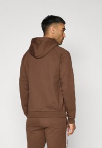 Sudadera con capucha marrón, con ribete en el dobladillo y en los puños. Presenta un detalle de logo en la manga y una tela suave y texturizada.