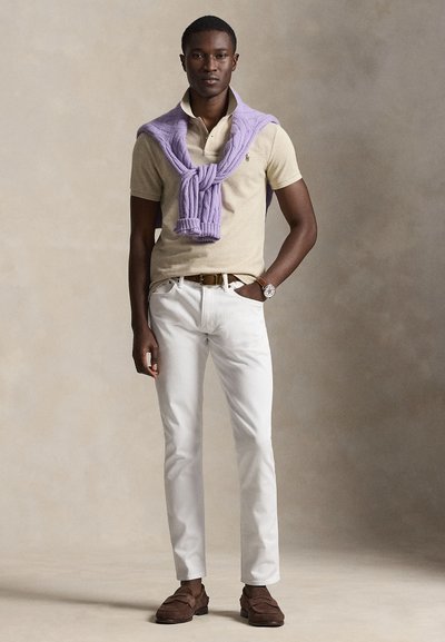 Moški model stoji z roko v žepu, oblečen bež polo majico, bele hlače, rjave loaferje, uro in sivkasto jopico, preloženo čez ramena.