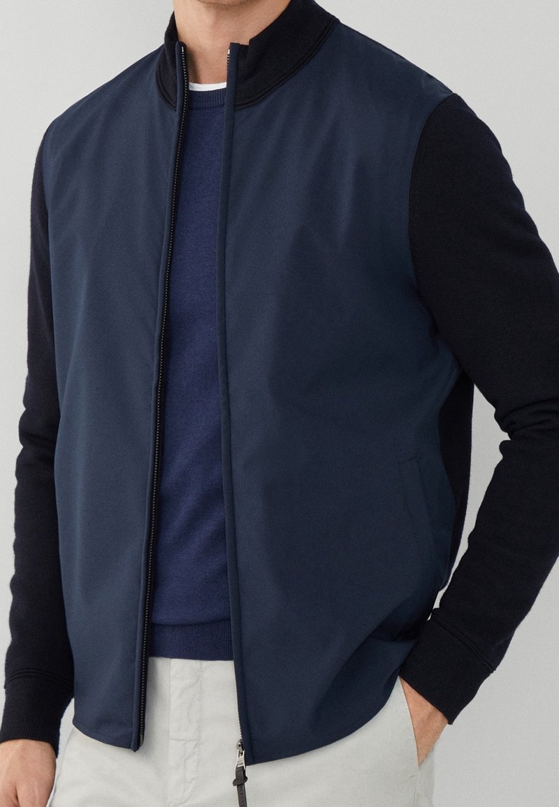 Homme portant une veste bomber bleu marine avec des manches noires, une chemise bleue en dessous et un pantalon clair, debout avec une main dans la poche.
