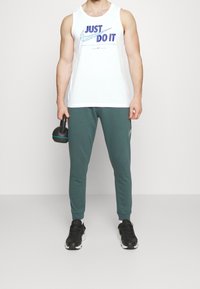 Man in een witte tanktop met de tekst "Just Do It", groene joggers, zwarte sneakers, en houdt een zwarte kettlebell in zijn rechterhand.