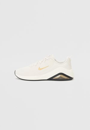 Witte sportschoen met gouden Nike-swoosh, gedempte zool met zwarte accenten en zichtbare luchtunit aan de hielzijde.