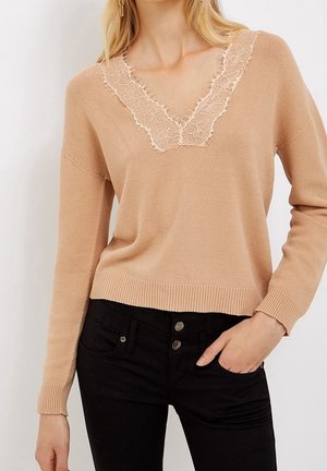 Kvinde iført en beige V-hals sweater med blondekanter og sorte højtaljede jeans, hånd i lommen, mod en ensartet lys baggrund.