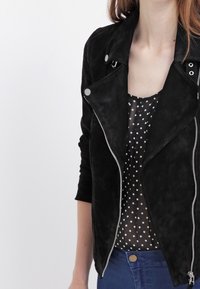 Chaqueta de motorista de ante negro con cremallera asimétrica, solapas y mangas largas, superpuesta sobre una blusa de gasa con lunares.