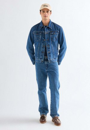 Młody mężczyzna w niebieskiej dżinsowej kurtce i dżinsach, brązowych butach oraz beżowej czapce baseballowej stoi na białym tle.