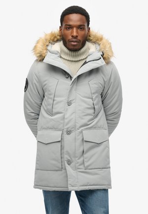 Parka gris claro con capucha con borde de piel, botones en el frente y grandes bolsillos. Llevada sobre un jersey de cuello alto color crema y jeans azules.