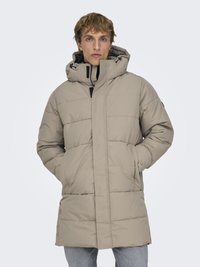 Giovane uomo che indossa un cappotto invernale trapuntato beige con cappuccio e mani nelle tasche, in piedi davanti a uno sfondo chiaro uniforme.