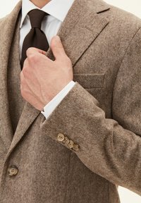 Brauner Blazer mit Fischgrätmuster aus strukturiertem Tweed, mit Reverskragen, vier runden Knöpfen am Ärmel und einer Brusttasche.