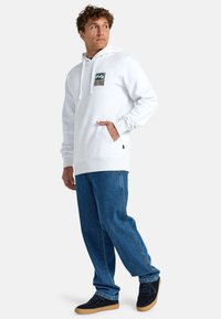 Light gray hooded sweatshirt with front pocket and graphic detail, paired with loose blue denim jeans and black shoes with tan soles.